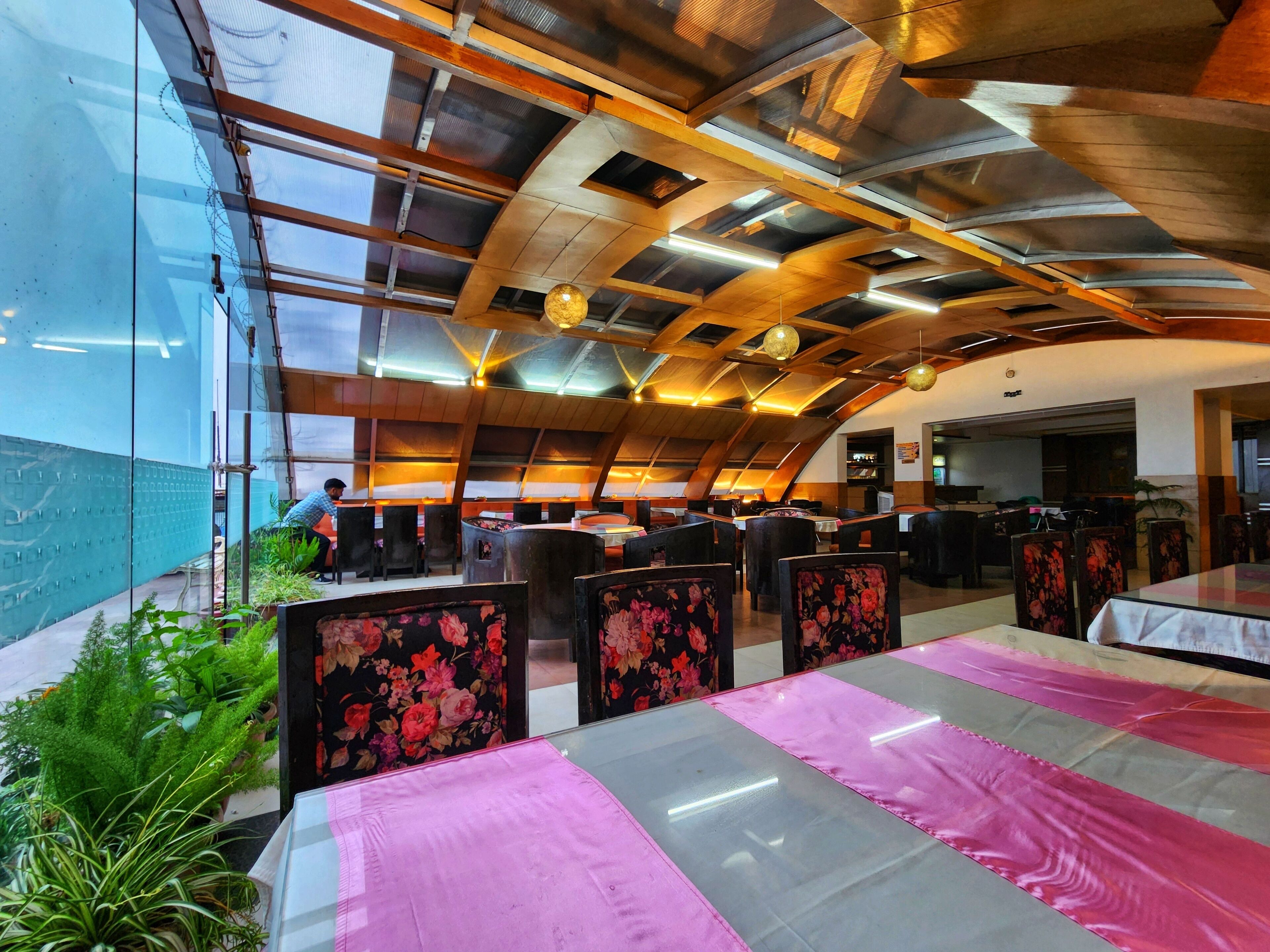 A restaurant with a modern design, featuring a high ceiling with exposed beams and a glass wall on one side. Tables are arranged with floral-patterned chairs.