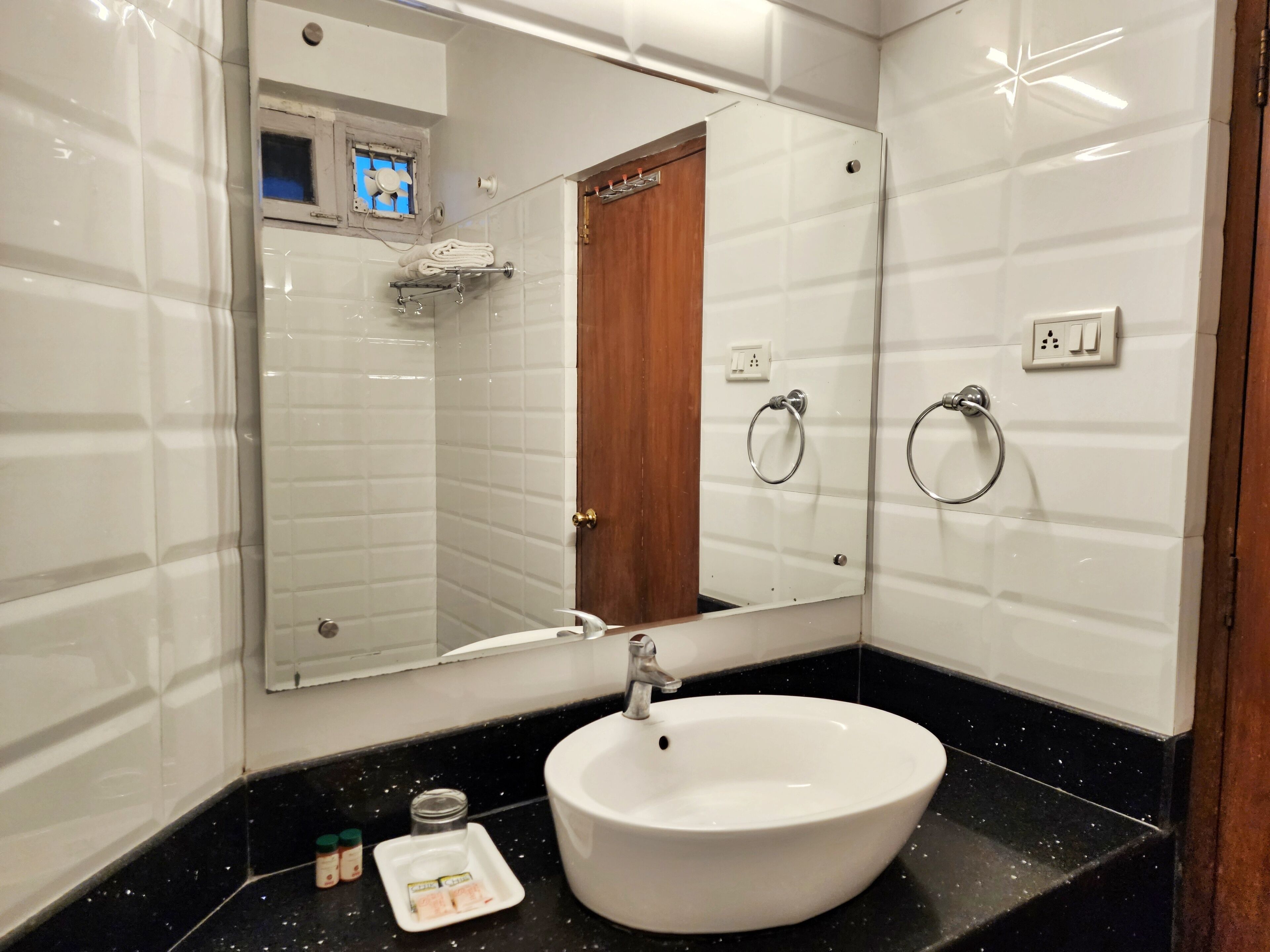 A bathroom with a white sink, black countertop, and a mirror above it. There is a towel rack and a door visible in the reflection.