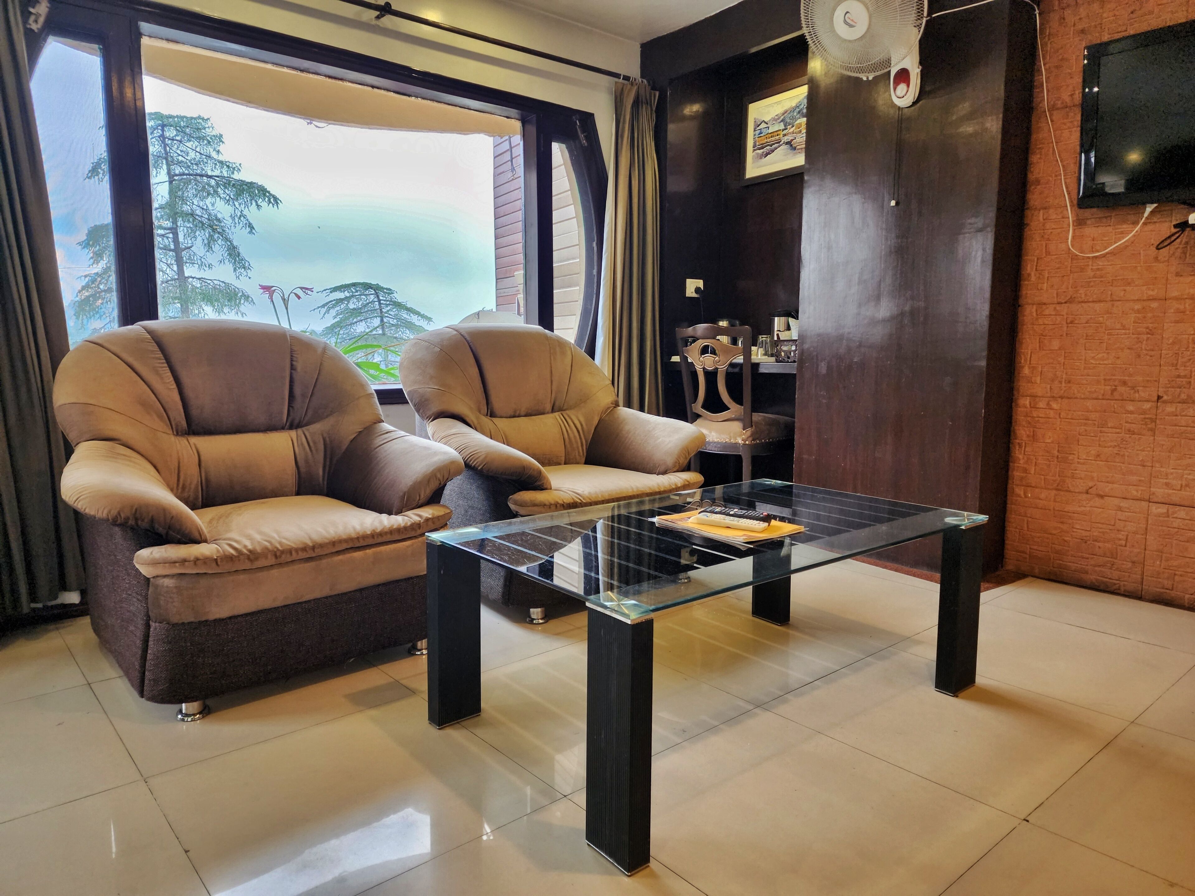 Un salon avec deux fauteuils en cuir marron, une table basse en verre, un téléviseur fixé au mur et une vue sur les arbres par une grande fenêtre.