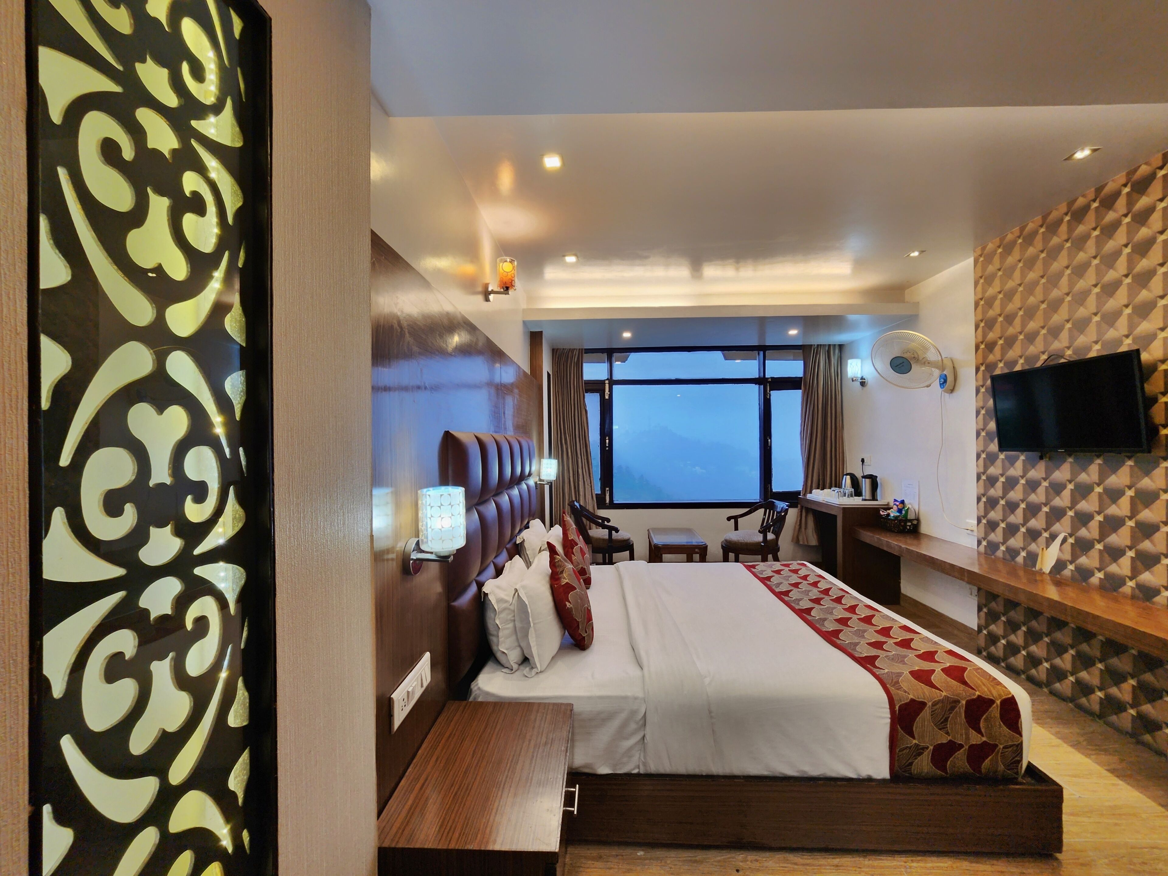 A hotel room with a bed, bedside table, lamp, TV, and a window with a view.