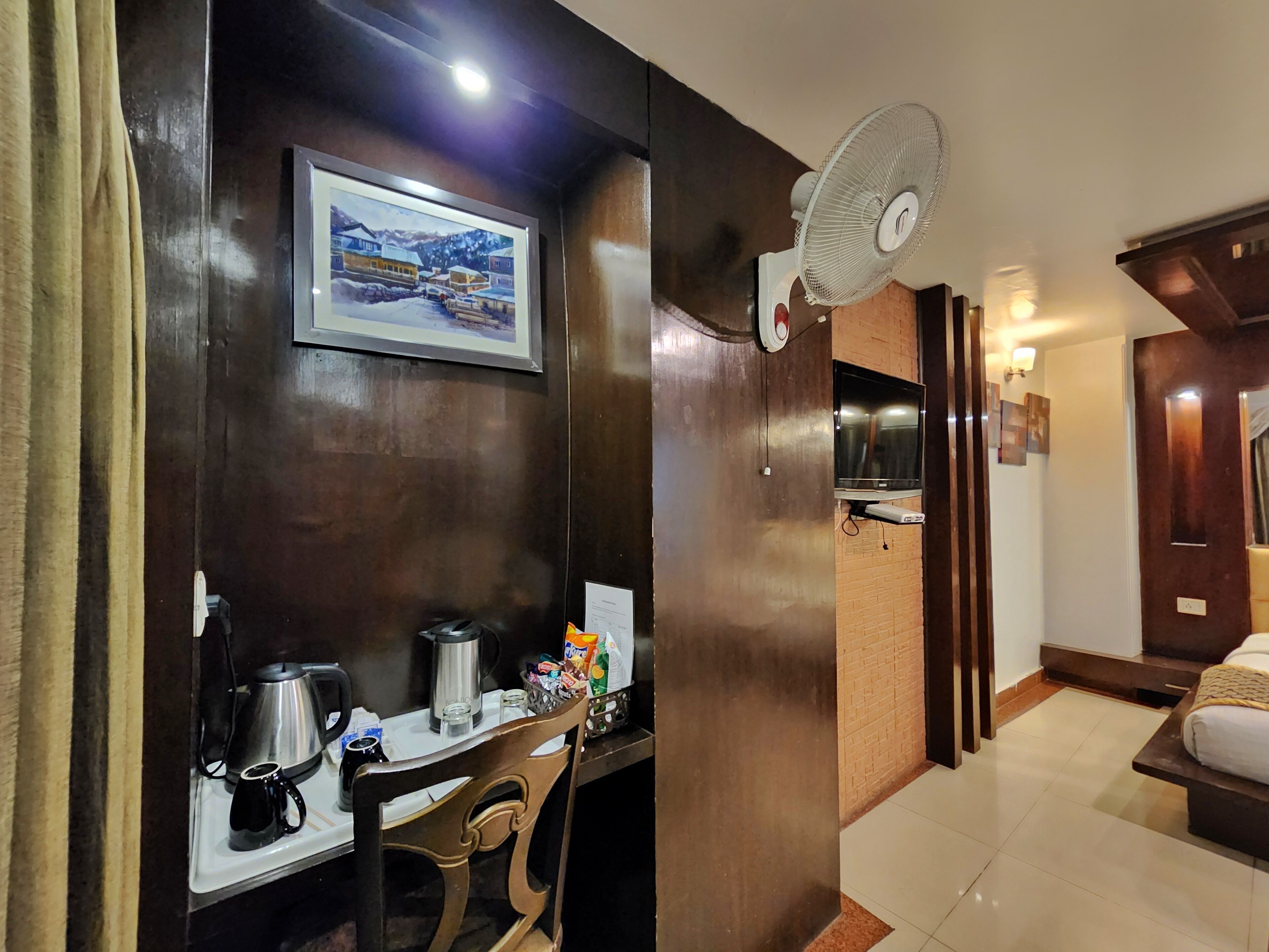 Une chambre d’hôtel équipée d’un bureau en bois, d’une chaise, d’une machine à café, d’une bouilloire, d’une petite table avec des en-cas, d’un téléviseur fixé au mur et d’un ventilateur sur pied.