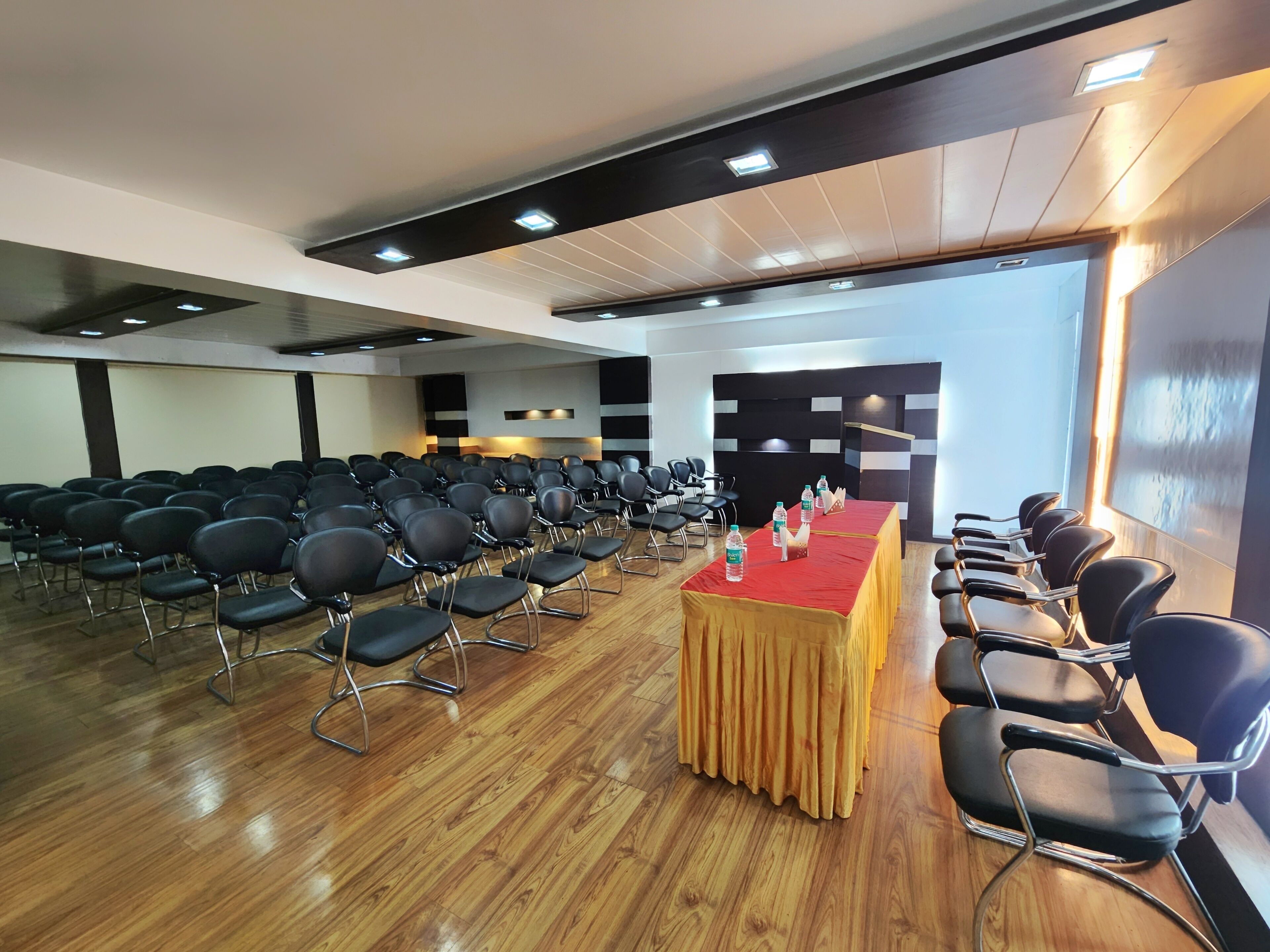 A conference room with rows of black chairs, a wooden table with water bottles, and a stage with a black backdrop.
