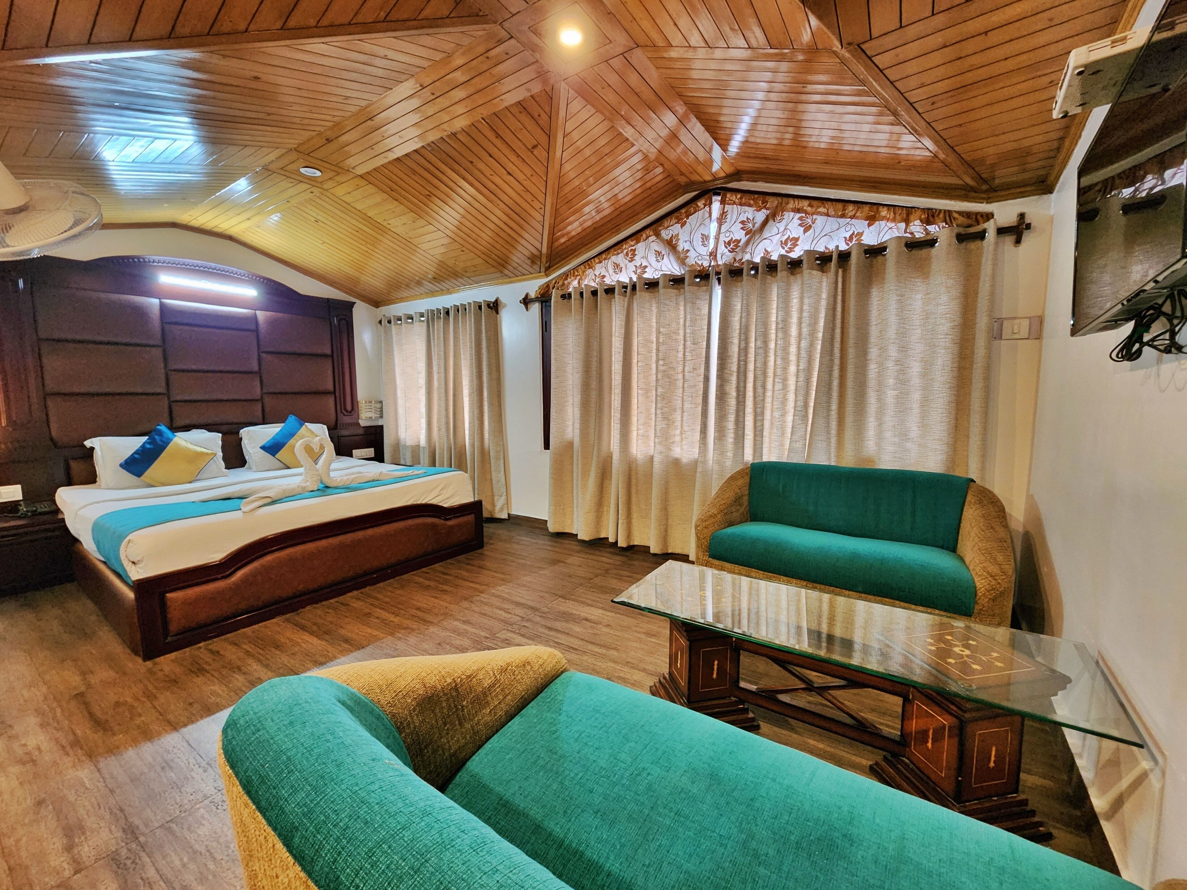 Una habitación de hotel con techo de madera, una cama con almohadas azules y amarillas, un sofá verde y una mesa de centro de vidrio.