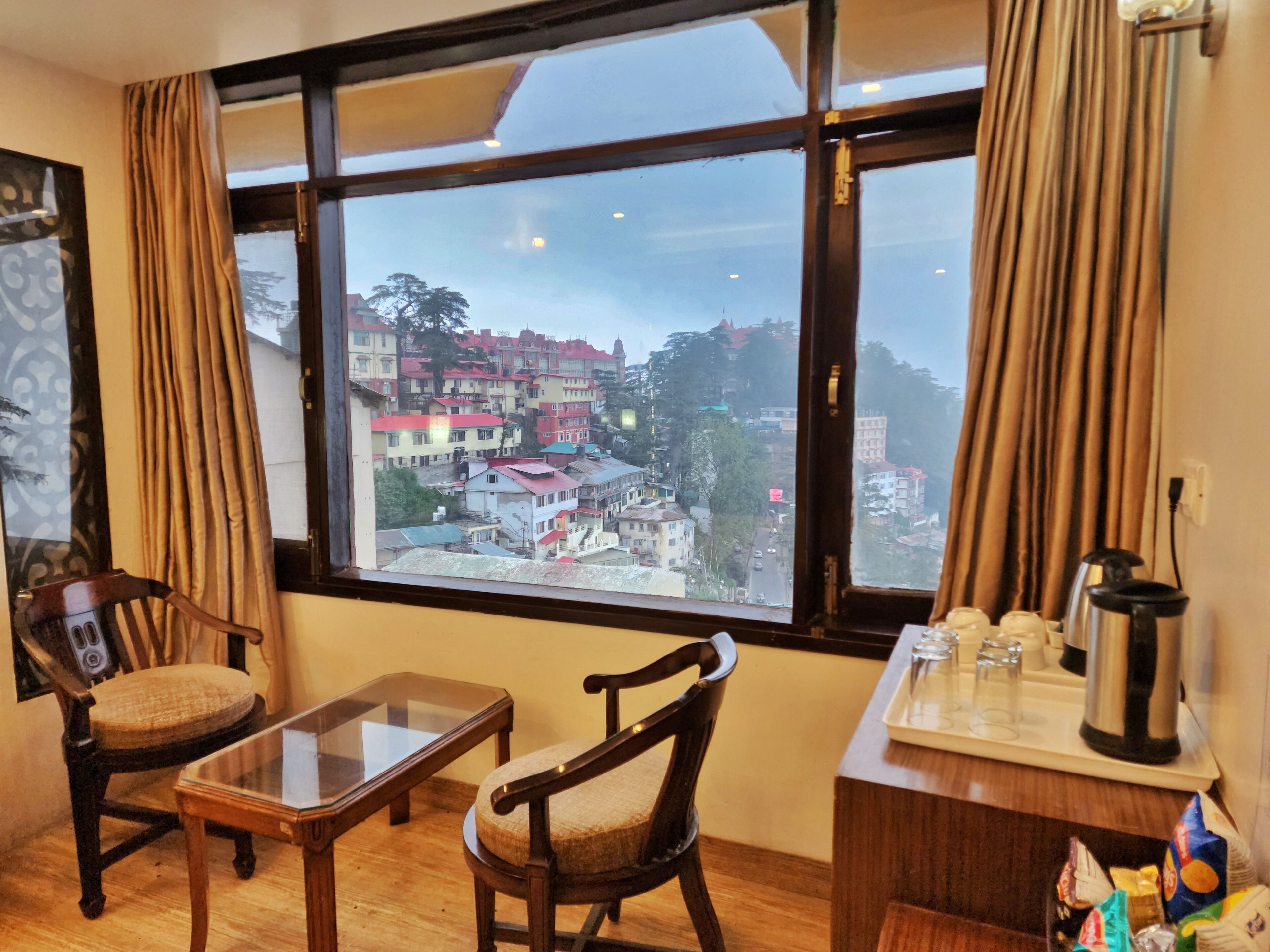 A hotel room with a large window offering a view of a cityscape. There is a wooden chair, a small table, and a coffee station with cups and a kettle.