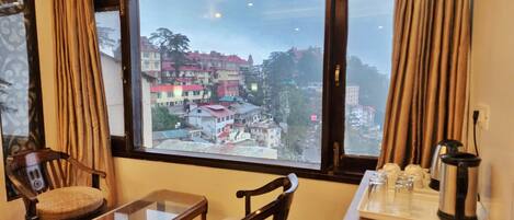 Ein Hotelzimmer mit großem Fenster und Blick auf die Stadt. Es gibt einen Holzstuhl, einen kleinen Tisch sowie eine Kaffeestation mit Tassen und Wasserkocher.