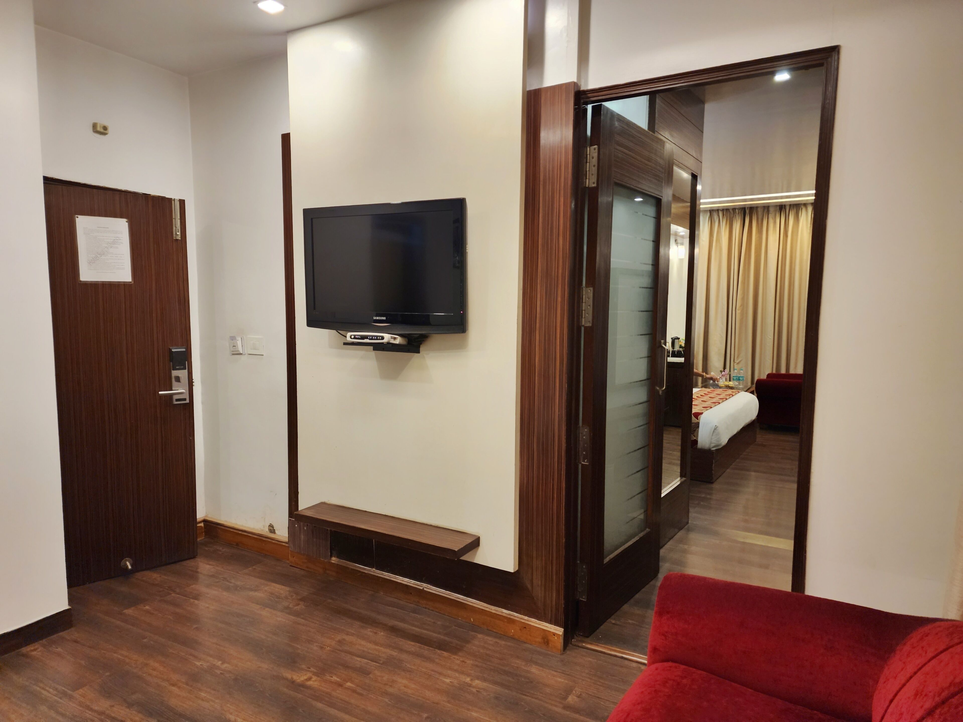 A hotel room with a wooden floor, a mounted TV, a door with a lock, and a mirror reflecting the room.