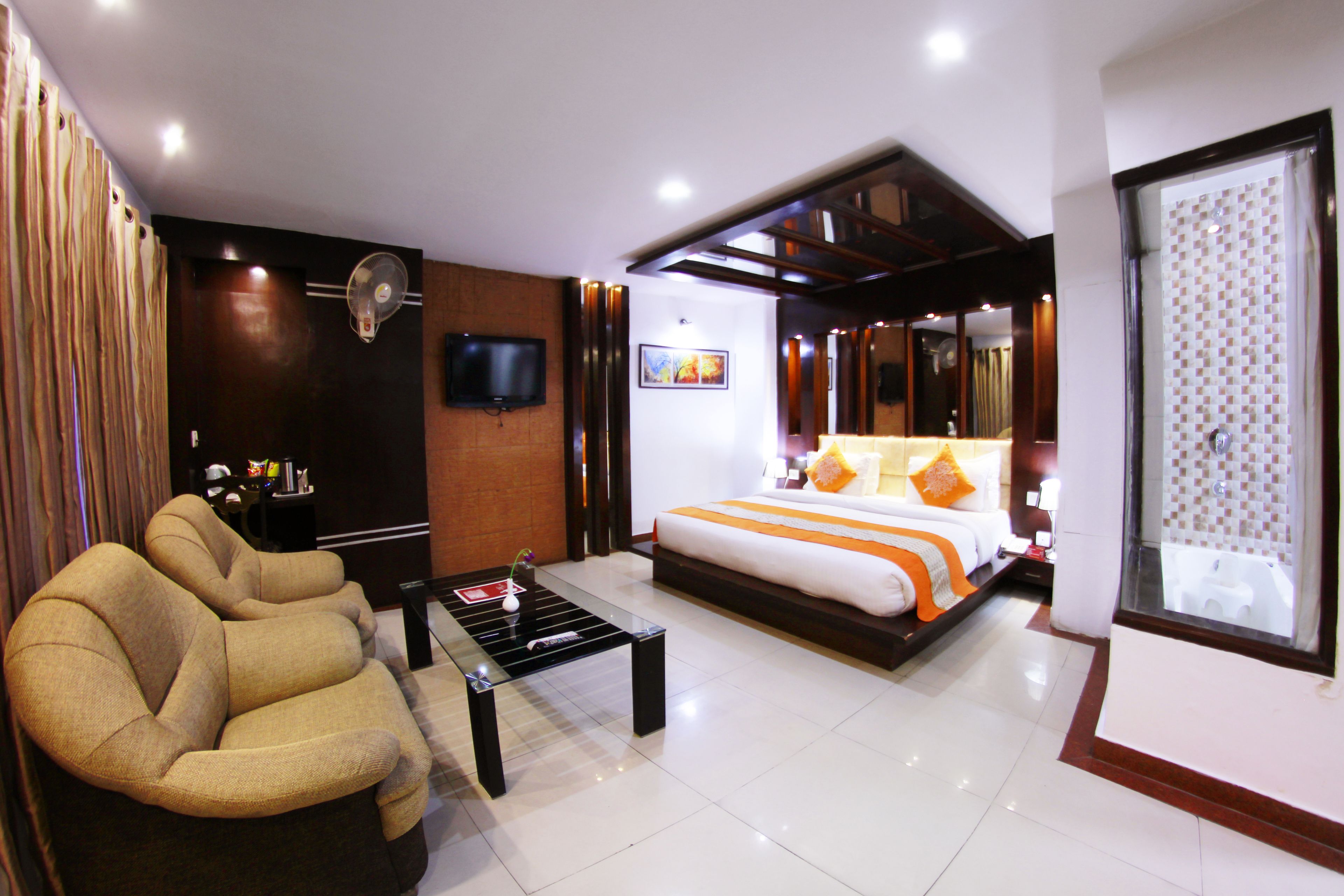 A modern hotel room with a large bed, a sofa, a coffee table, a TV, and a bathroom with a shower and toilet.