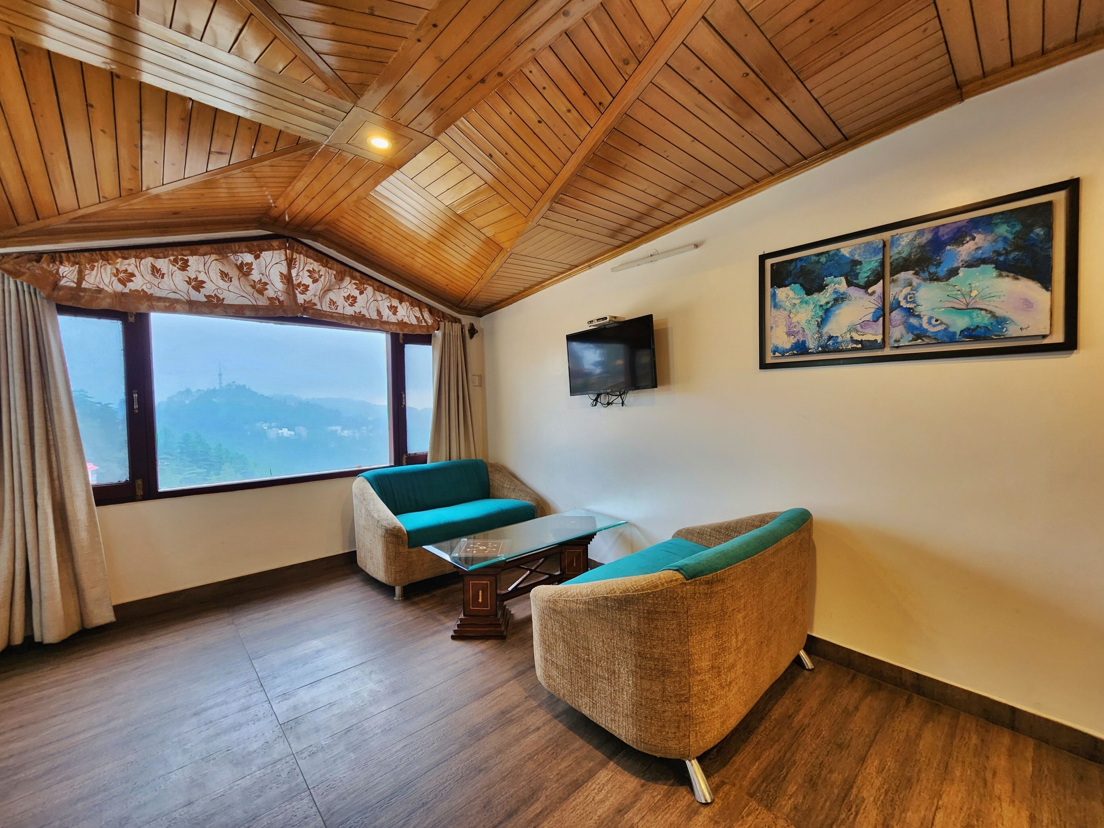 A cozy room with wooden ceiling beams, a large window with a view, two sofas, a TV, and abstract wall art.