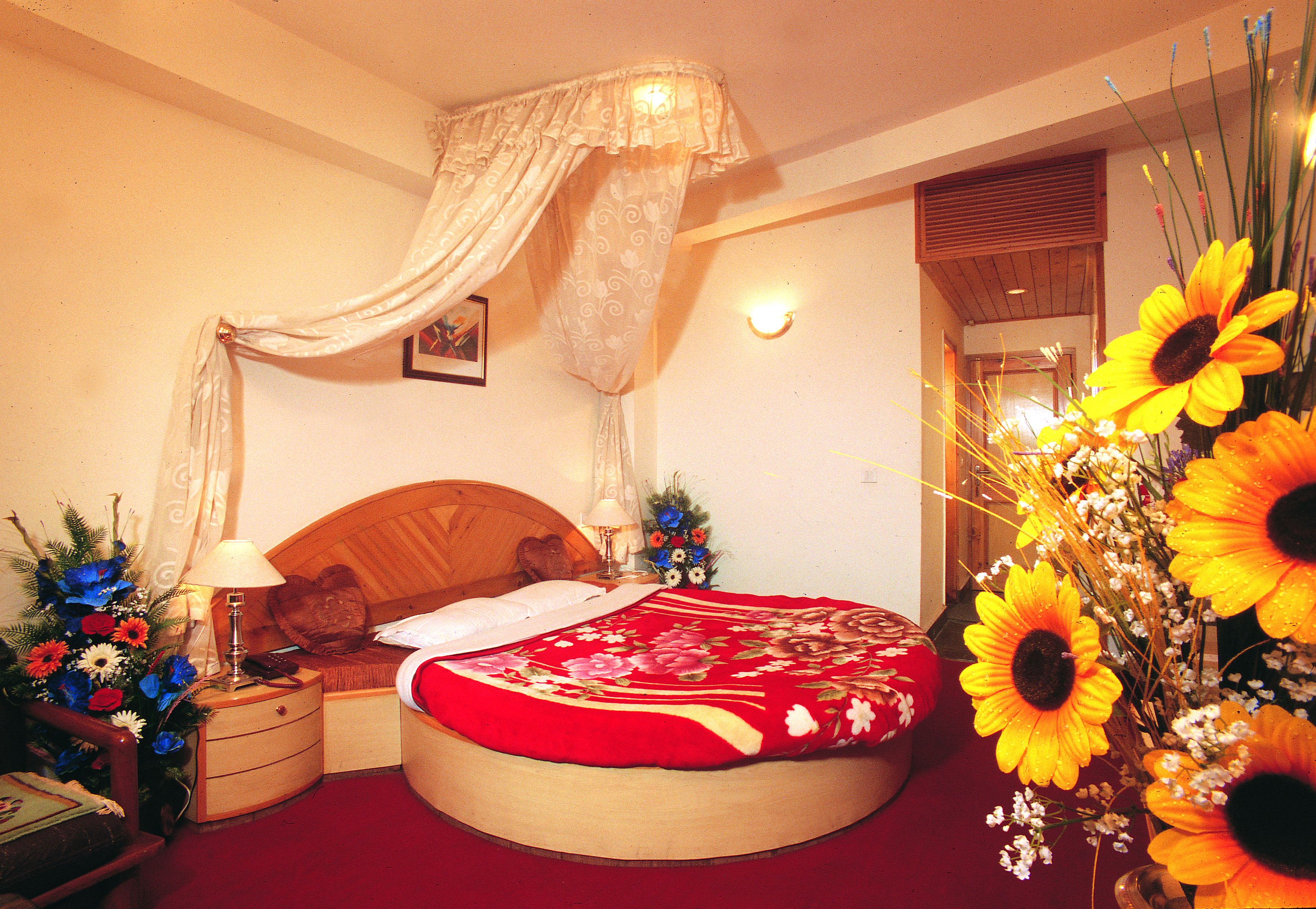 A round bed with a red floral bedspread, a wooden headboard, and a nightstand with a lamp and a vase of flowers.