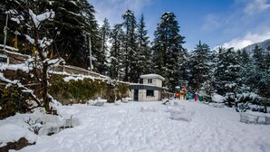 Área externa coberta de neve, com bancos e um pequeno edifício, rodeada por altas árvores perenes.