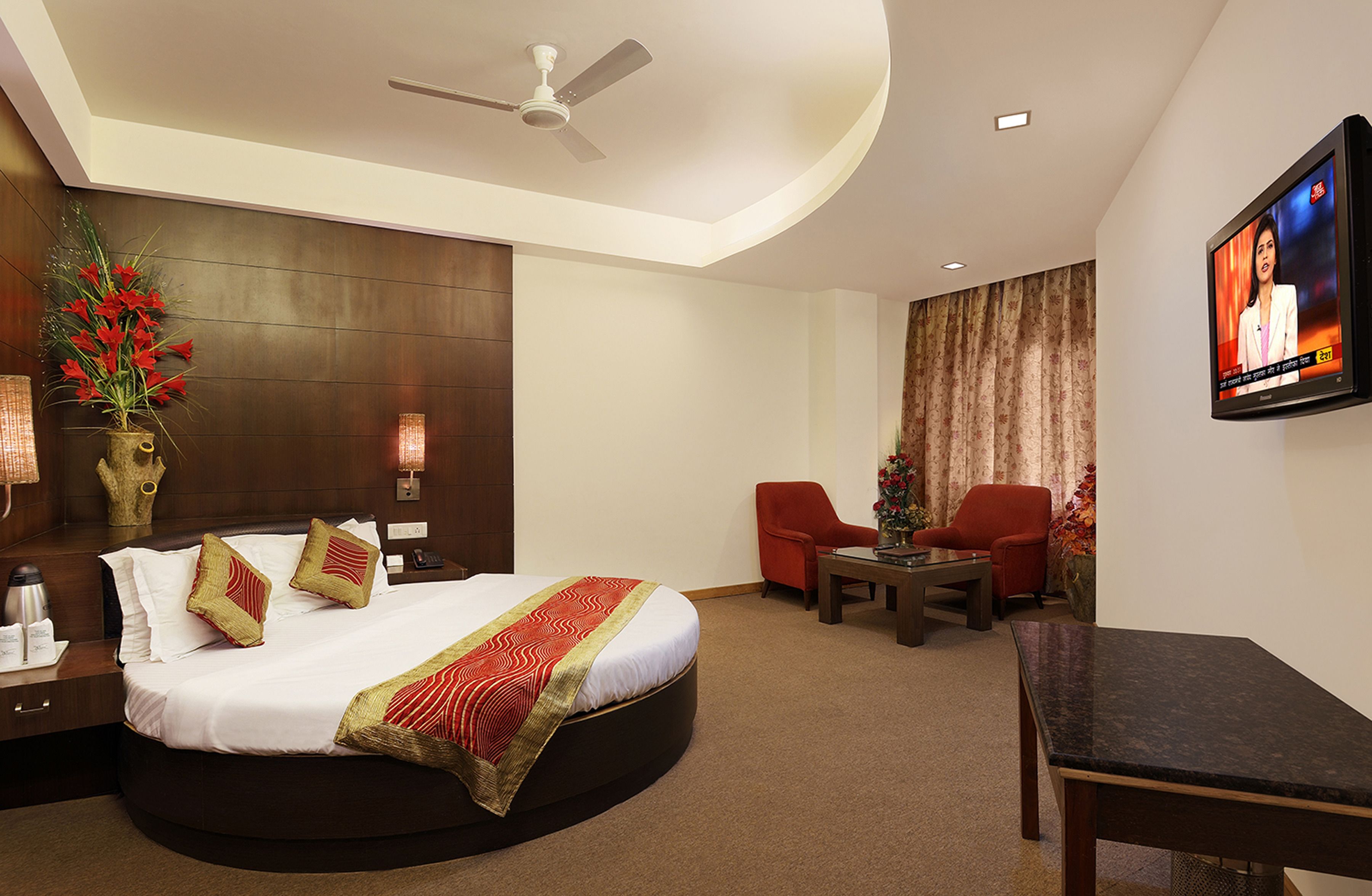 A hotel room with a large bed, a desk, two chairs, a TV, and a ceiling fan.