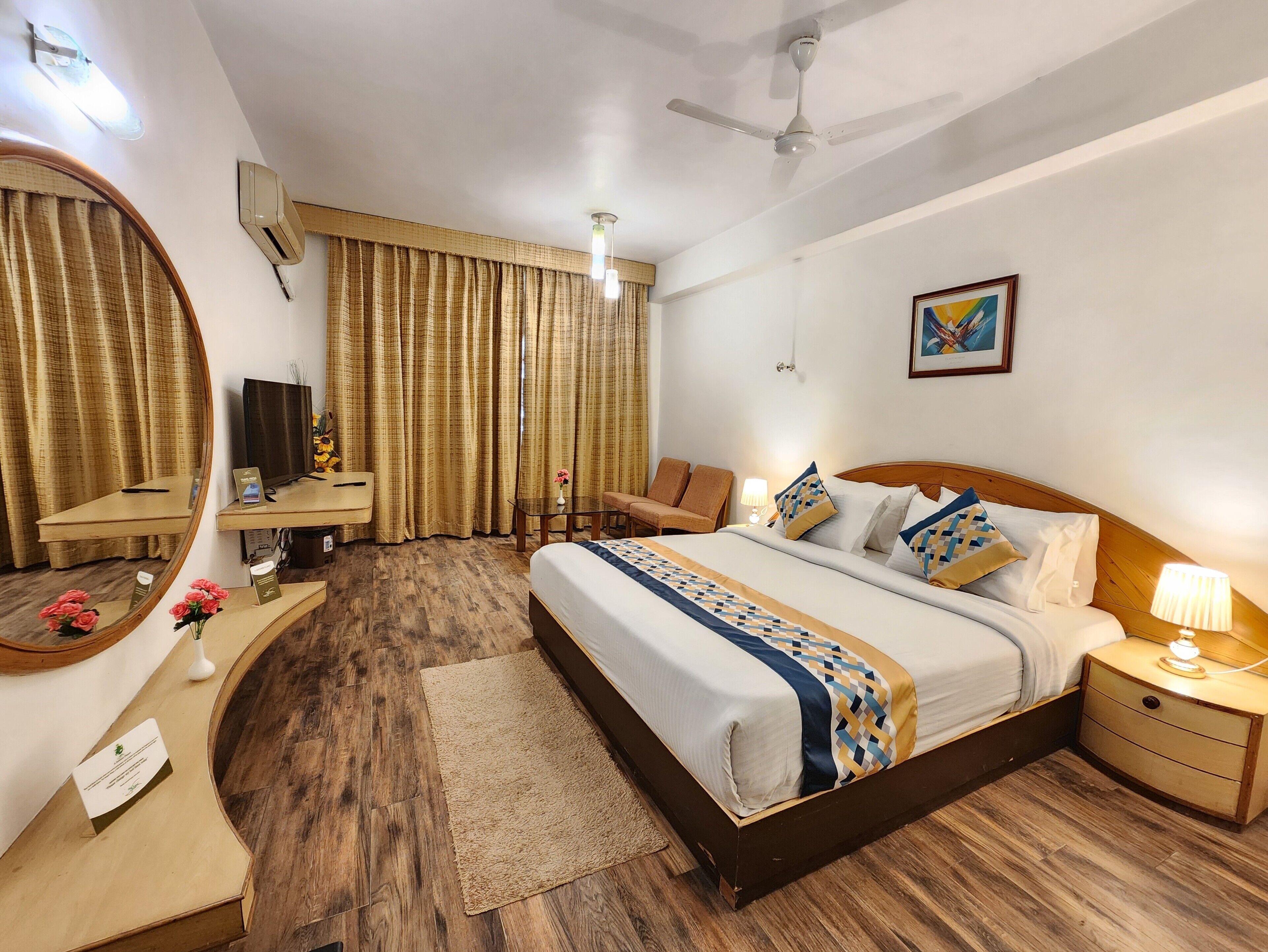 A hotel room with a bed, bedside table, lamp, chair, desk, mirror, curtains, and a ceiling fan.