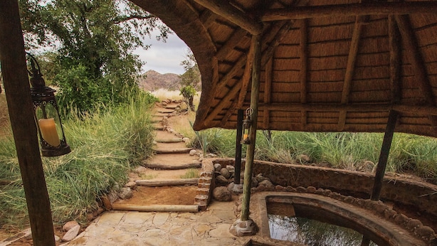 Un sendero de piedra lleva a una construcción rústica de madera con techo de paja, una pequeña piscina y un farol.