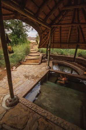 A stone pathway leading to a rustic wooden structure with a thatched roof, a small pool, and a lantern post. - Huab Lodge (Kamanjab)