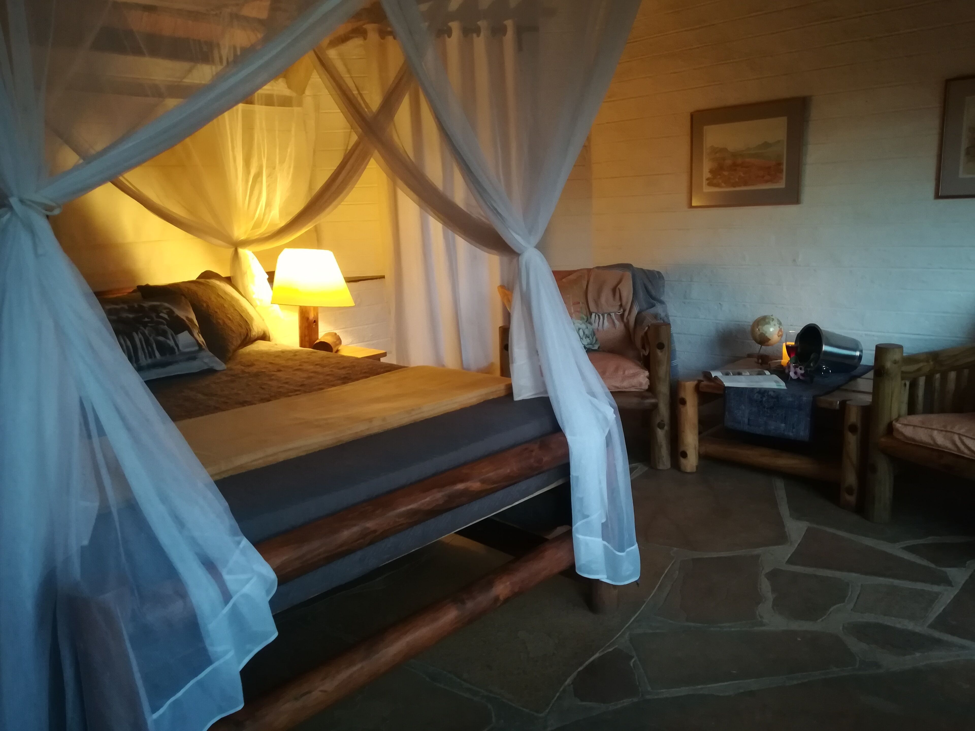Una habitación acogedora con una cama bajo dosel, una mesita de noche con lámpara, una silla de madera y suelo de piedra.