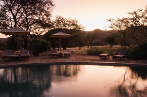 Zona junto a la piscina con sillones de descanso y sombrillas, rodeada de árboles y colinas.
