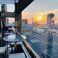 Un bar en la azotea con barandilla de vidrio, mesas y sillas, con vista a un paisaje urbano al atardecer.