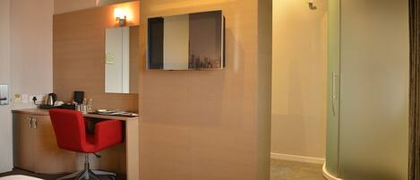 A hotel room with a bed, a desk with a chair, a mirror, a wardrobe, and a shower.
