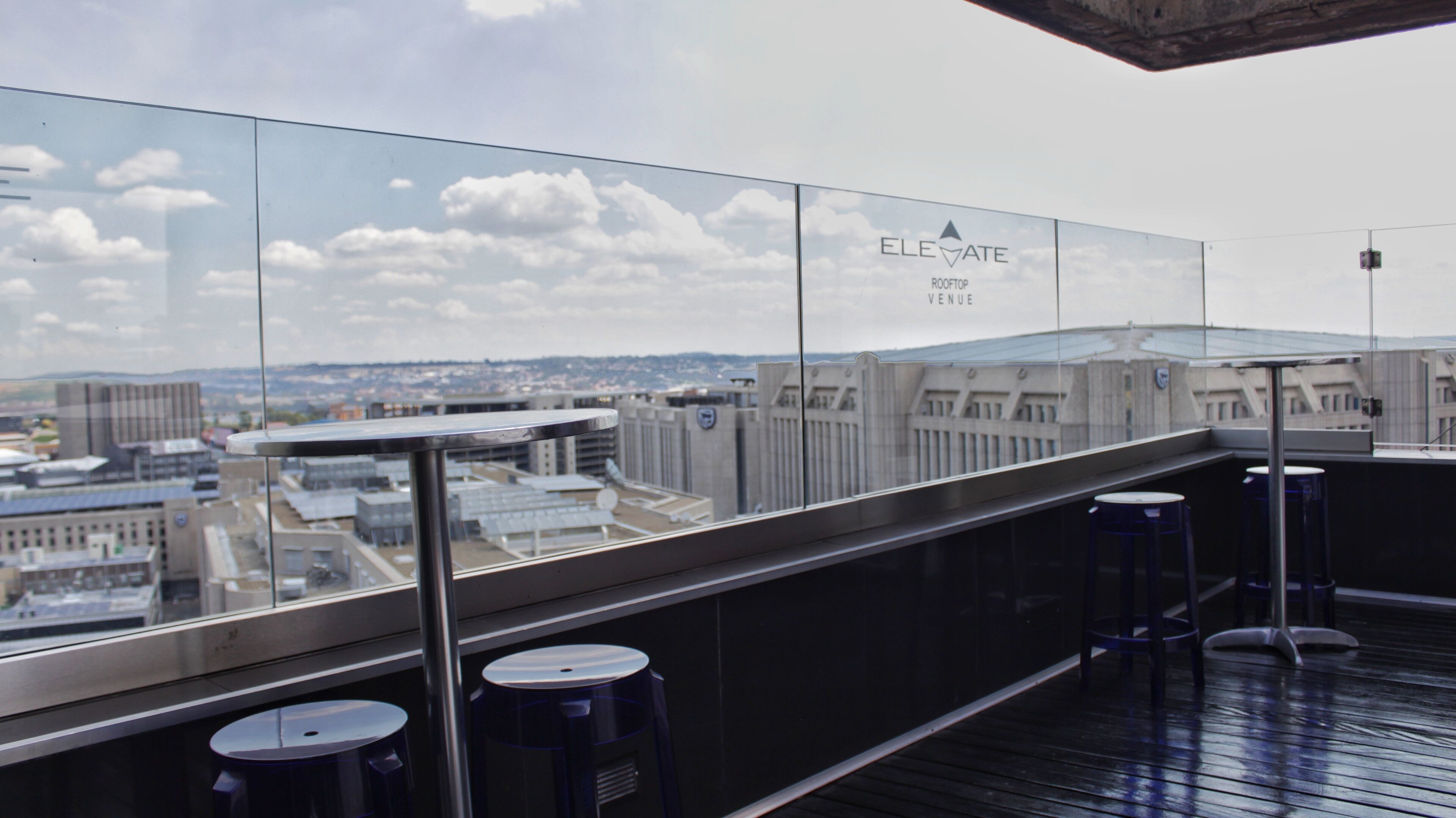 Un bar en la azotea con barandilla de vidrio, que ofrece vistas panorámicas de la ciudad y un cartel que dice "ELEVATE ROOFTOP VENUE".