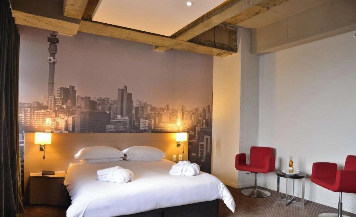 Ein Hotelzimmer mit einem Bett, zwei Nachttischlampen, einem Stadtbild-Wandgemälde und zwei roten Stühlen.