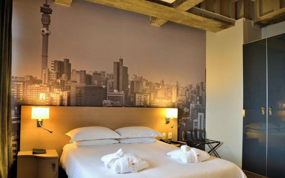 A hotel room with a bed, two bedside lamps, a cityscape mural, a wardrobe, and a desk with a chair.