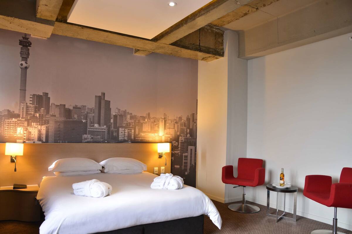 A hotel room with a bed, two red chairs, a cityscape mural, and a city view.