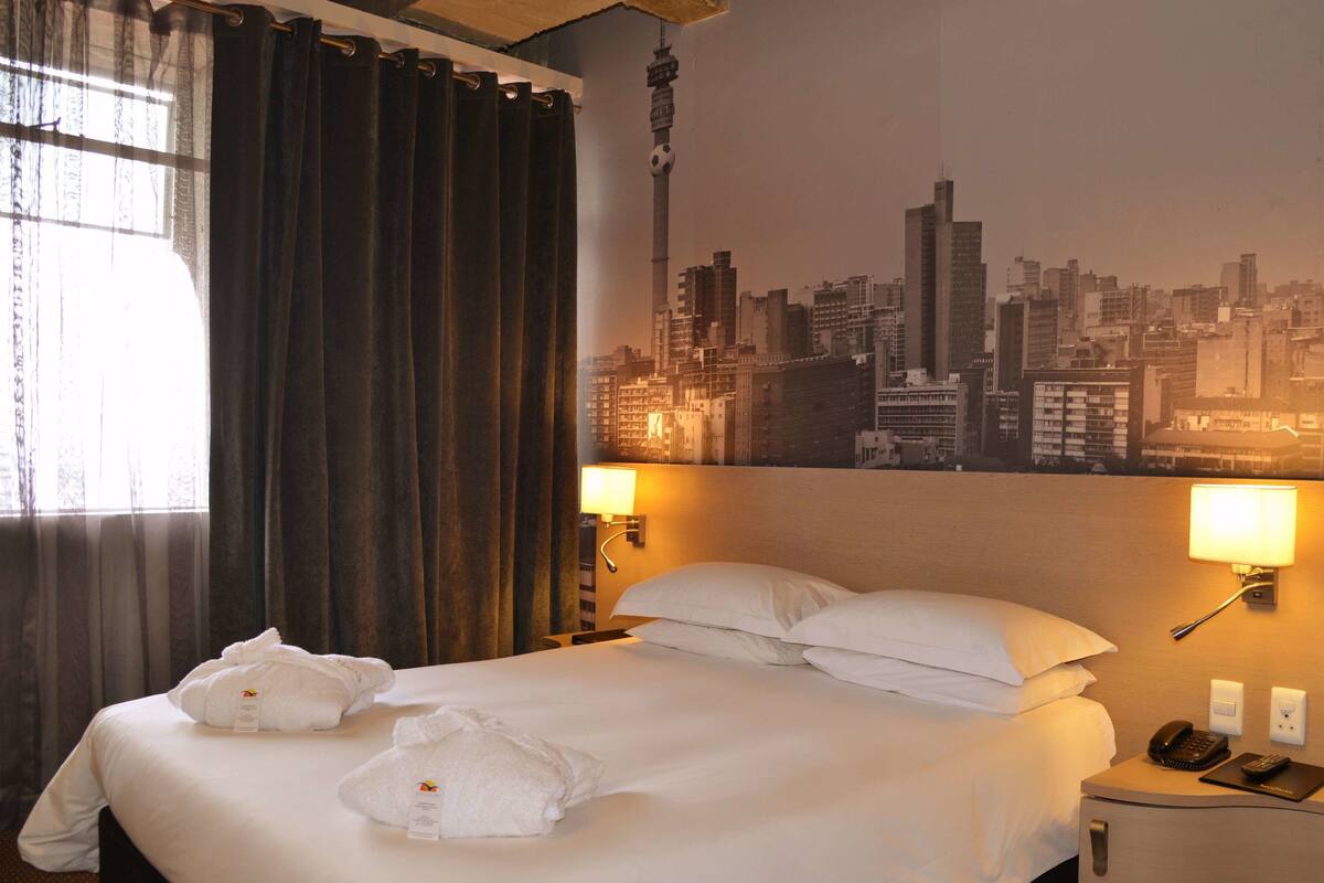 A hotel room with a bed, two bedside lamps, a cityscape wall mural, and a window with curtains.