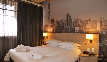 Una habitación de hotel con una cama, dos lámparas de noche, un mural de un paisaje urbano y una ventana con cortinas.