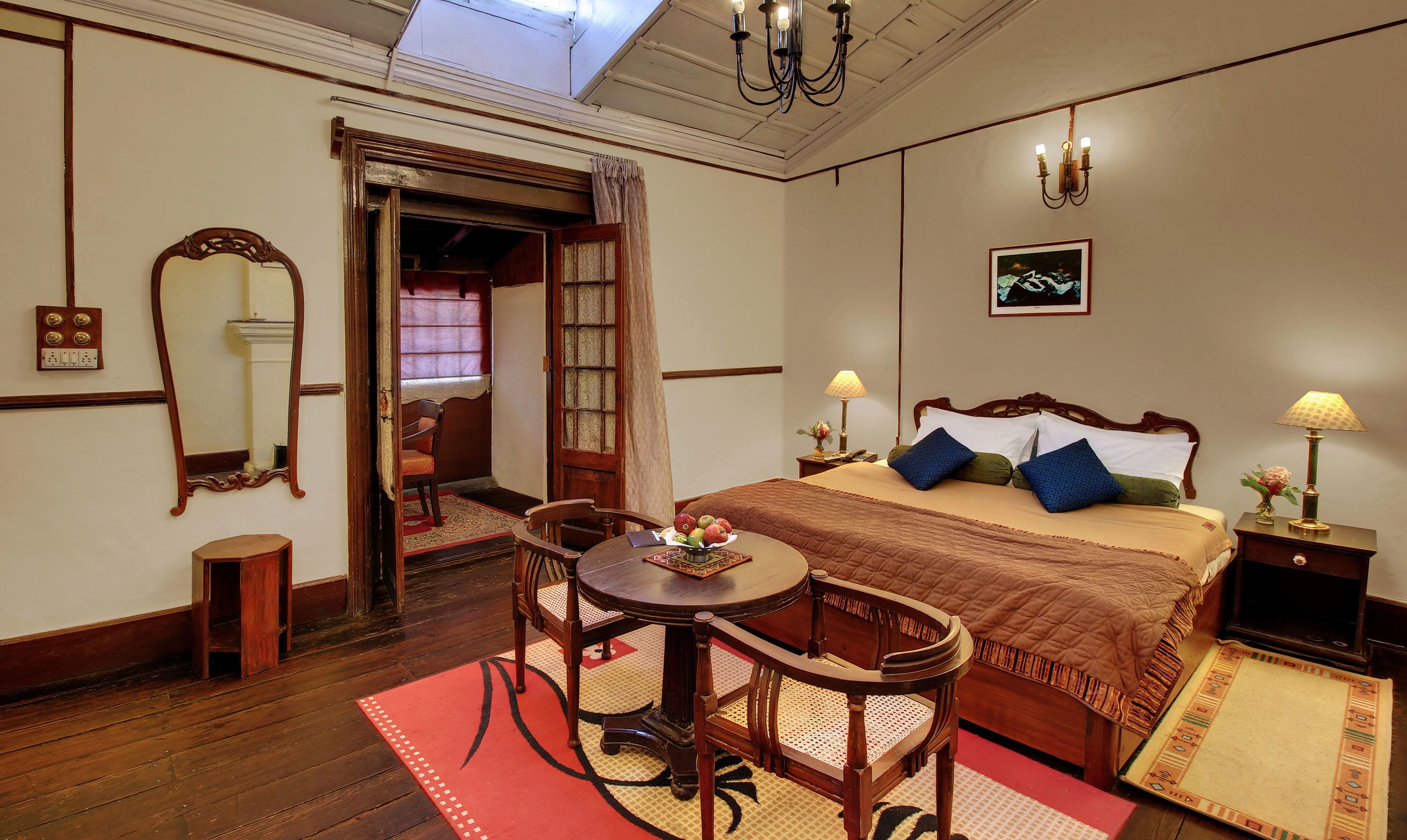 Una habitación tradicional con cama, mesa y silla. Hay un espejo en la pared, una araña y un cuadro.