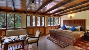 A wooden-paneled bedroom with a large bed, a chair, a small table with cups and a remote control, and large windows offering a forest view.