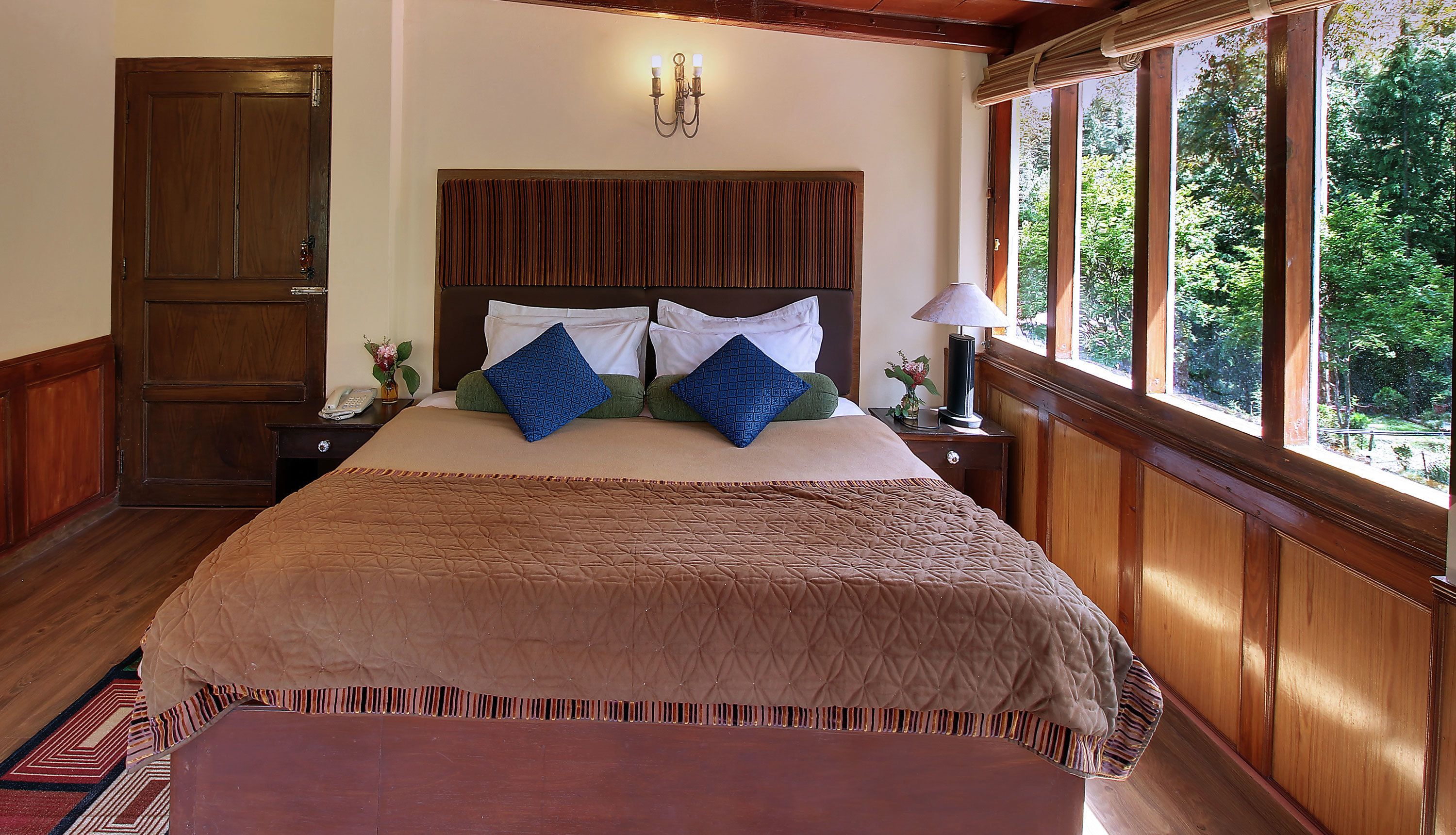 A large bed with a quilted cover, two bedside tables with lamps, a wooden headboard, a wooden door, and a window with a view of greenery.