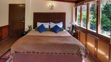 A large bed with a quilted cover, two bedside tables with lamps, a wooden headboard, a wooden door, and a window with a view of greenery.