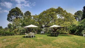 A grassy outdoor area with two white tables and chairs, surrounded by trees and a clear sky.