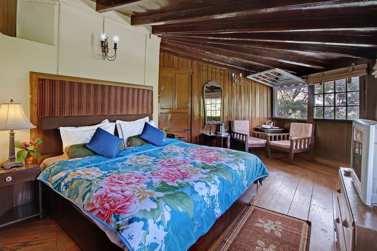 A bedroom with a large bed, a nightstand, a lamp, a mirror, and a view of the outdoors through a window.