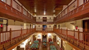 Un vestíbulo de hotel con varios niveles, barandillas de madera, una escalera central y múltiples mesas de comedor.