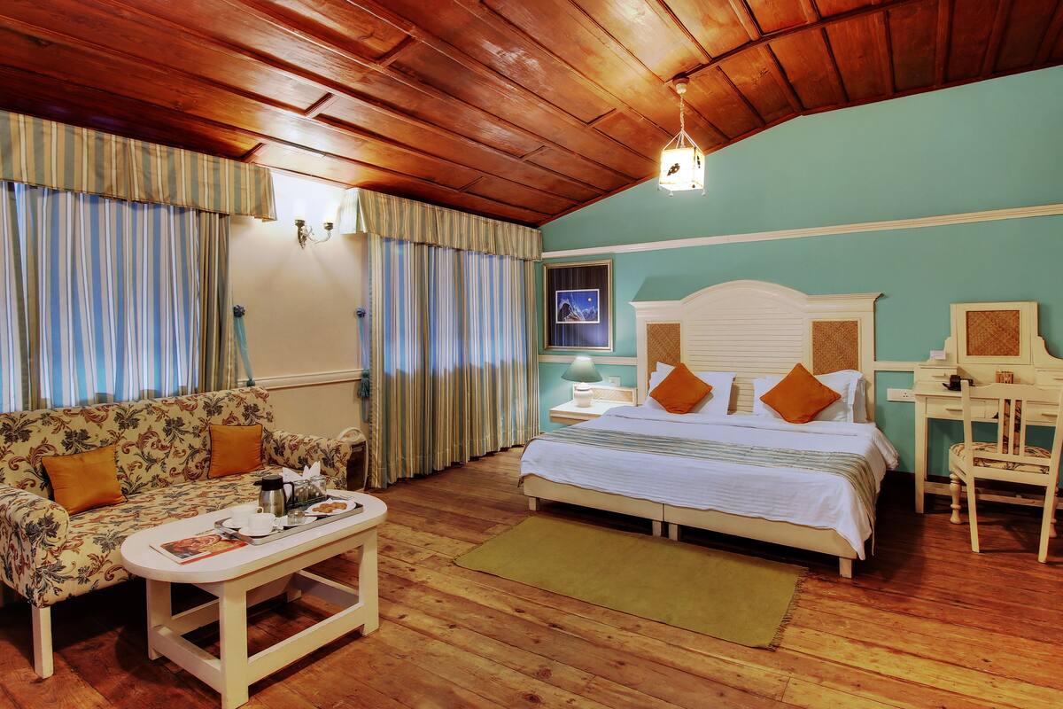A cozy room with a wooden ceiling, a bed with white bedding and orange pillows, a floral sofa, a small table with a tray of breakfast items, and a desk with a chair.