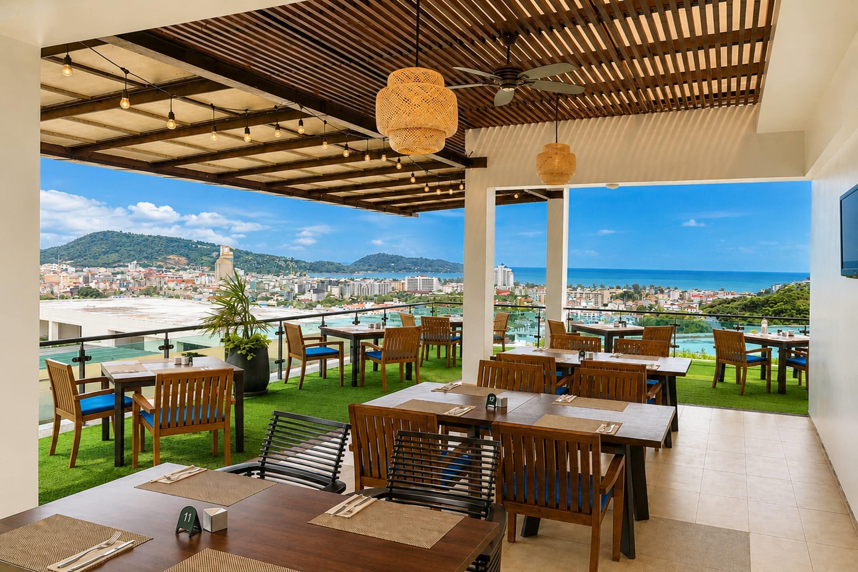 Un área de comedor al aire libre con mesas y sillas, vistas a la ciudad y al océano, y un techo con vigas a la vista y luces colgantes.