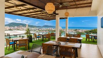 Un área de comedor al aire libre con mesas y sillas, vistas a la ciudad y al océano, y un techo con vigas a la vista y luces colgantes.