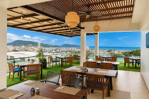 An outdoor dining area with tables and chairs, a view of a cityscape and ocean, and a ceiling with exposed beams and hanging lights.