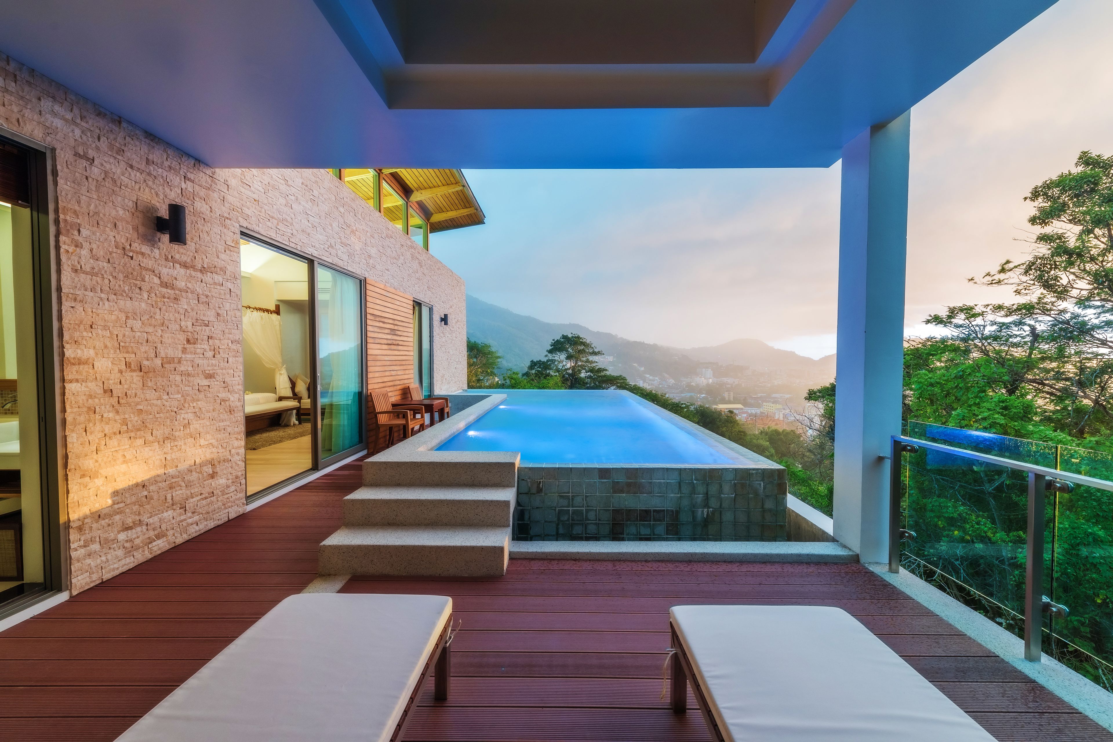 A modern outdoor pool area with a wooden deck, lounge chairs, and a view of mountains.
