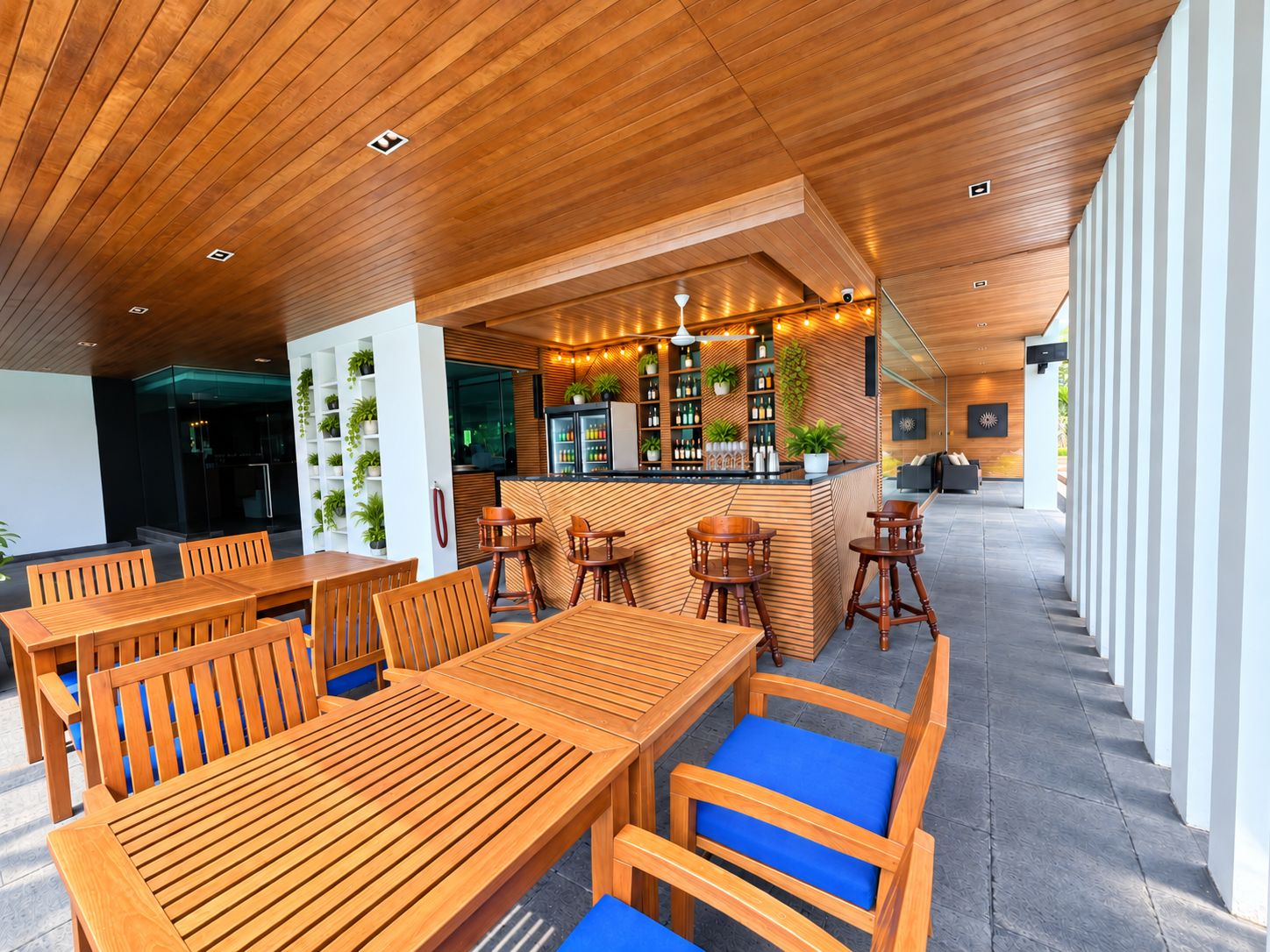 Ein moderner Außenbereich zum Essen mit Holztischen und -stühlen, eine Bar mit Regalen voller Flaschen und Blick auf die Umgebung.