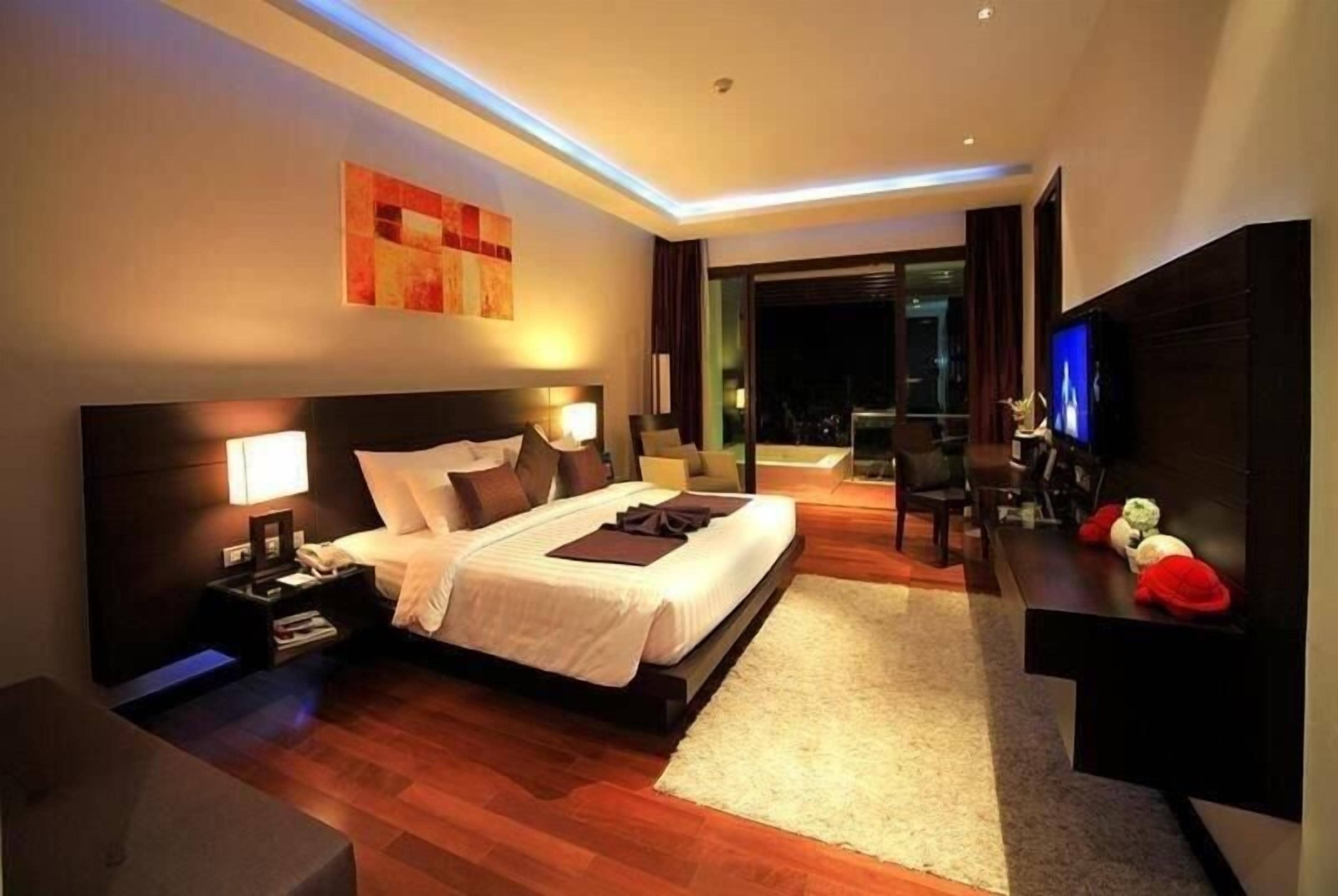 Una habitación de hotel moderna con una cama grande, mesitas de noche, un televisor de pantalla plana y vistas a la ciudad de noche.