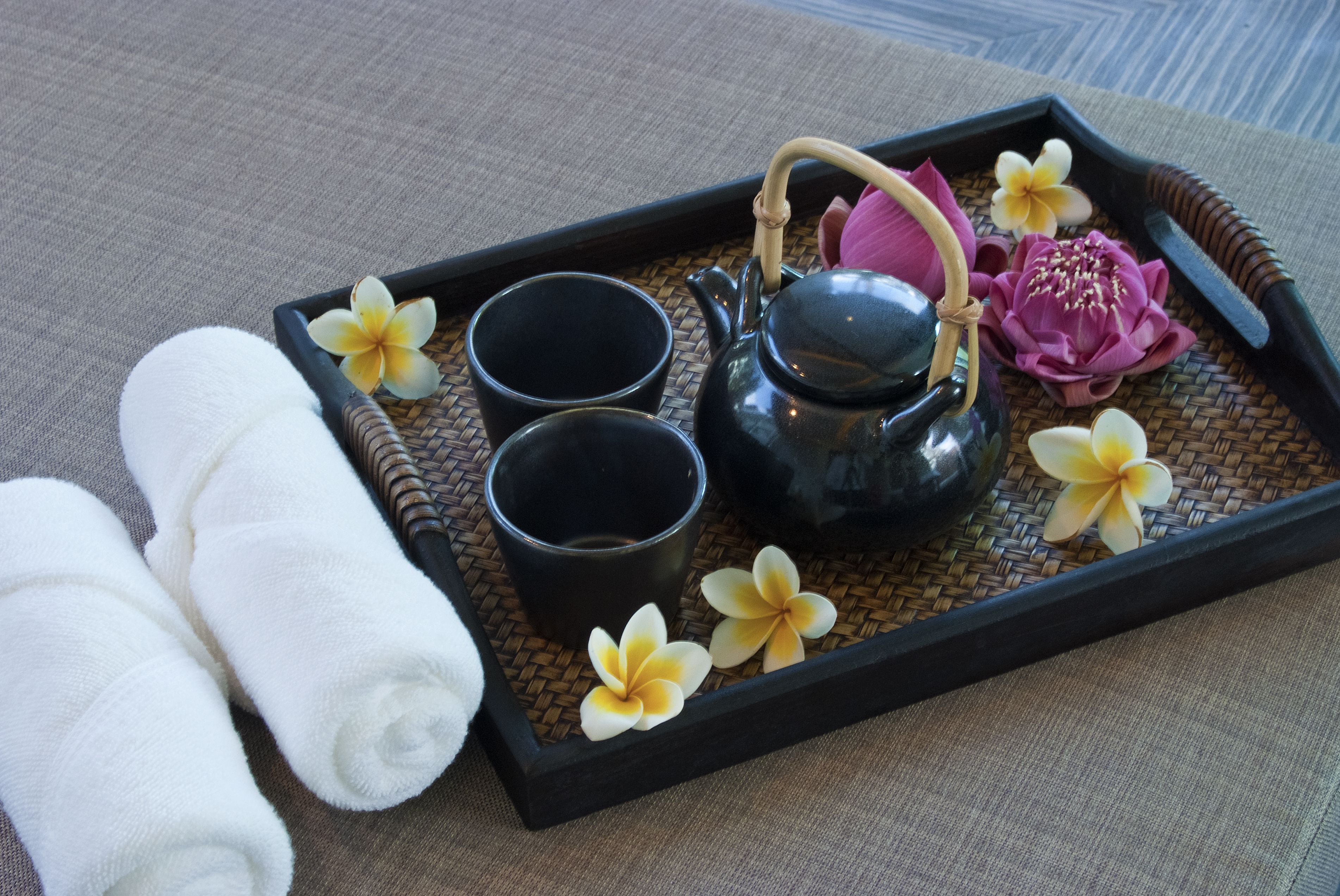 A tray with a black teapot, two black cups, and a pink flower on a woven mat.