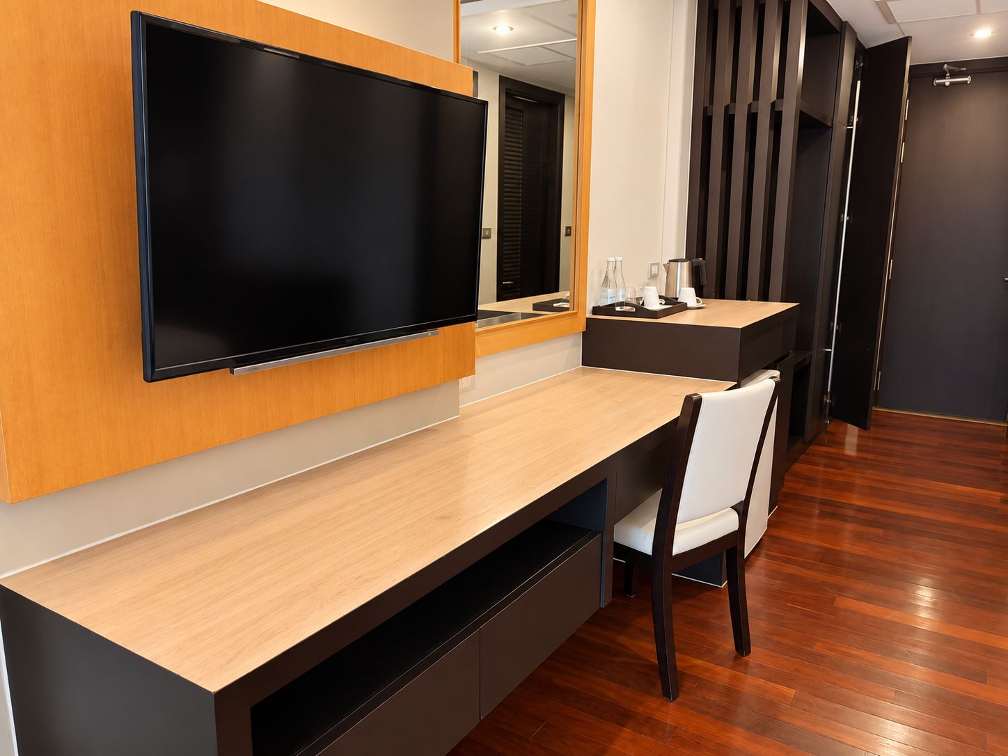 A modern hotel room with a large flat-screen TV mounted on the wall, a wooden desk with a chair, and a small coffee table.