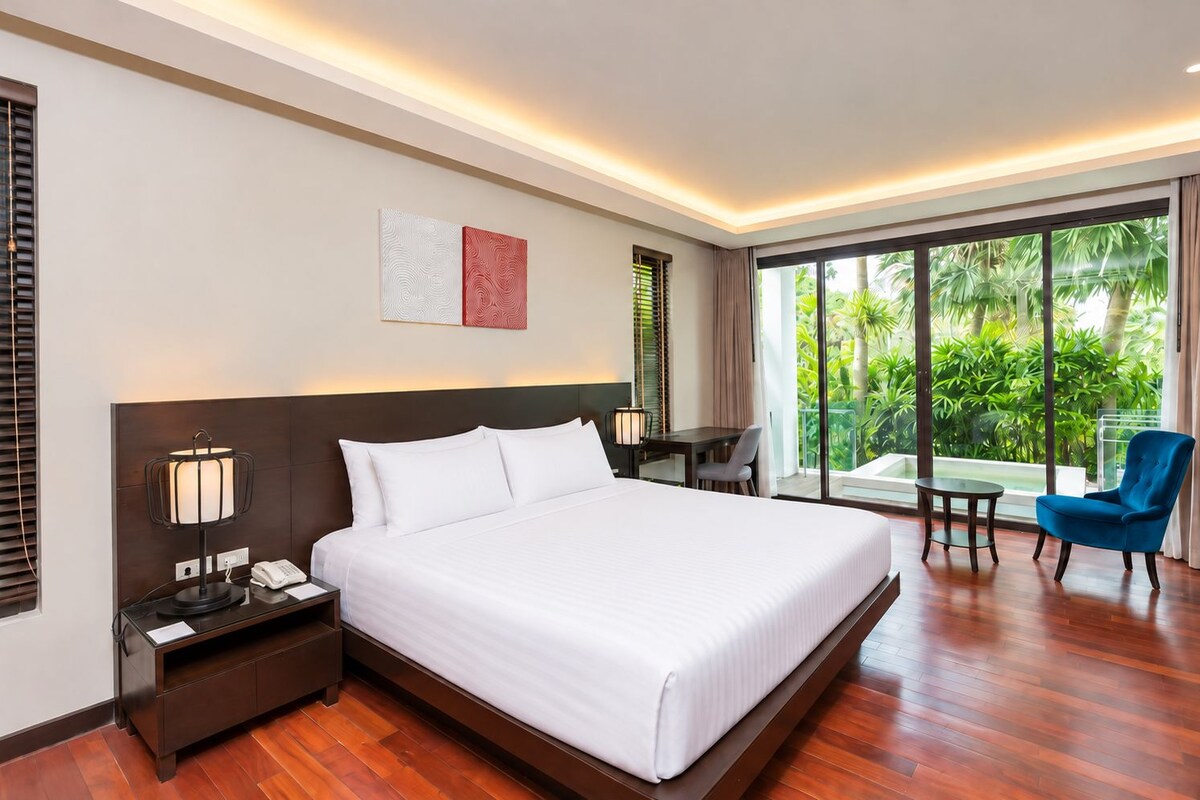 Habitación de hotel con una cama grande, mesita de noche con lámpara, escritorio, silla y vista a zonas verdes a través de amplios ventanales.