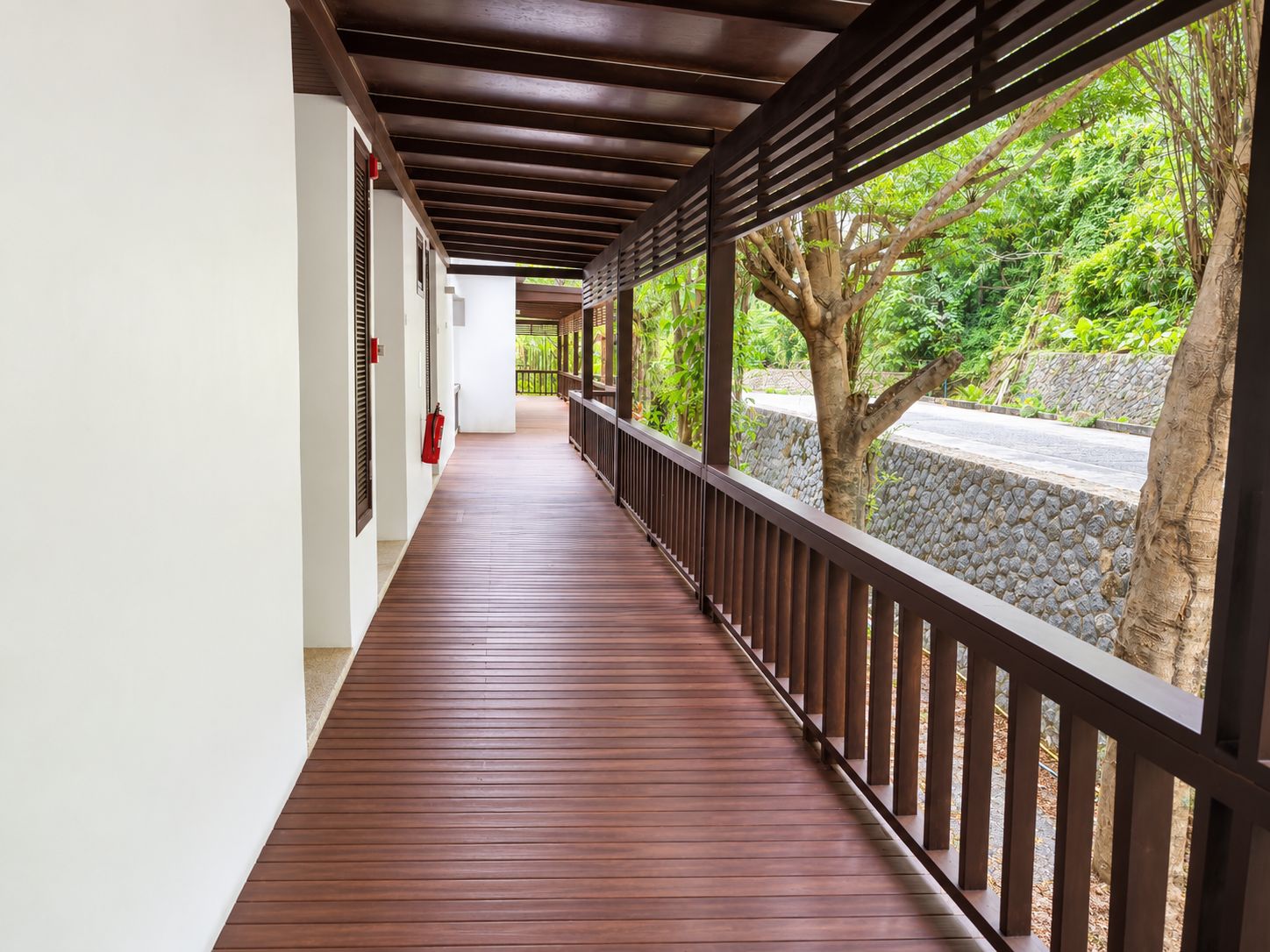 A covered corridor with a wooden floor and metal railing, leading to a stone wall and greenery.