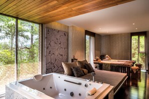 A modern bedroom with a large bathtub, a bed with multiple pillows, and a view of trees through large windows.