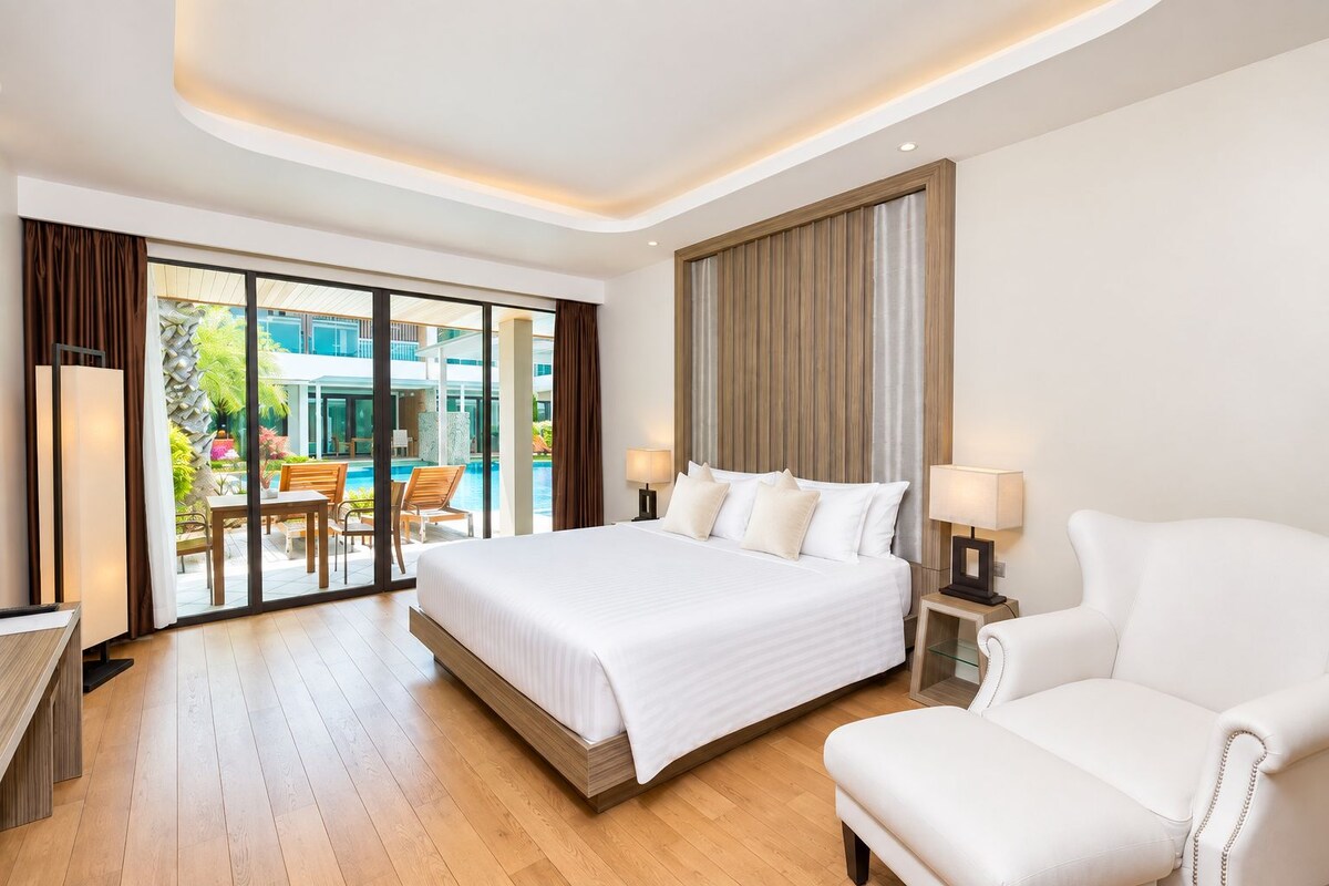 Habitación de hotel con una cama grande, una zona de estar con una silla, un escritorio y vistas a una piscina al aire libre.