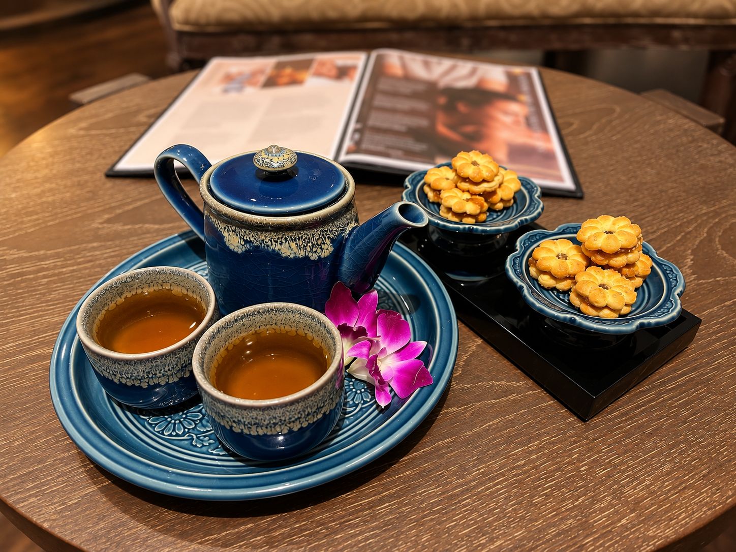 Uma mesa com um bule azul, duas xícaras de chá, um prato de biscoitos e uma flor rosa.