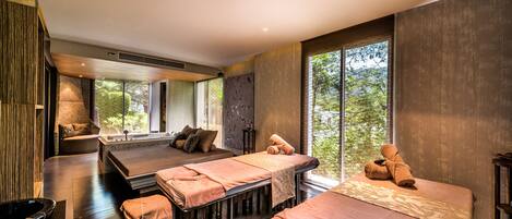 Une salle de spa équipée de deux tables de massage, d’une grande fenêtre donnant sur la forêt et d’un mur en pierre.